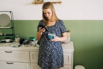 Junge werdende Frau in Zierkleidung mit Tasse Heißgetränk plaudert auf Handy im Haus — Stockfoto
