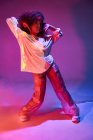 Молода афроамериканська танцівниця повністю вписується в світле неформальне вбрання, торкаючись кучерявого волосся і дивлячись на камеру під час танців у темній студії в неонові вогні. — стокове фото