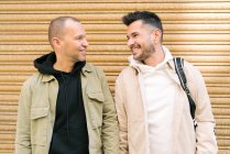Позитивные молодые многонациональные геи в стильной одежде, улыбающиеся и смотрящие друг на друга, держась за руки на городской улице — стоковое фото