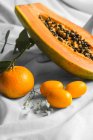 Half of pawpaw with seeds between tangerines and cumquats with leaves on white crumpled cloth — Foto stock