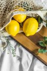 Bunte ganze Zitronen im Zero-Waste-Sack in der Nähe von welligem Pflanzenzweig auf Holzschneidebrett auf geknittertem Textil — Stockfoto