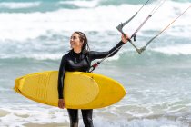 Female athlete in wetsuit with control bar looking away on sandy shore against foamy ocean after practicing kiteboarding — Stock Photo