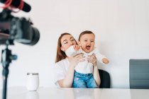 Joven madre con un lindo bebé en vueltas hablando y grabando video en la cámara para blog personal mientras está sentado en el escritorio en casa - foto de stock