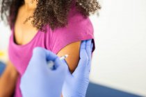 Especialista médica femenina irreconocible en uniforme protector y guantes de látex vacunando a una paciente afroamericana anónima en la clínica durante el brote de coronavirus - foto de stock