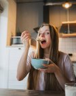 Молода жінка з ложкою і мискою насолоджується смачними кільцями кукурудзи, дивлячись на камеру на кухні — стокове фото