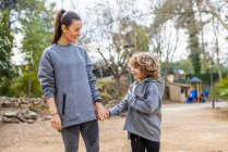 Mother in sportswear holding boy by hand while strolling on walkway and talking against trees looking at each other — Foto stock