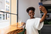 Feliz joven afroamericano femenino en blusa blanca tomando selfie en el teléfono móvil mientras está sentado en la mesa alta cerca de la pared de vidrio en la cafetería moderna - foto de stock