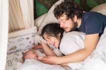 Сверху веселые родители, взаимодействующие с милым маленьким ребенком, лежа в постели дома — стоковое фото
