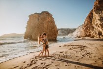 Hombre en forma de cuerpo completo abrazando joven novia atractiva en traje de baño mientras se enfrían juntos en la costa arenosa cerca de acantilados ásperos en el día soleado en Fyriplaka Milos - foto de stock