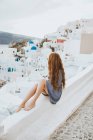 Обратный вид на неузнаваемую женщину-путешественницу, любующуюся деревней Ия на острове Санторини в ветреный день в Греции — стоковое фото