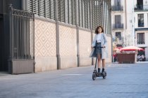 Повний вміст тіла африканка-американка молода жінка в повсякденному одязі їздить скутер на мощеному тротуарі і дивиться геть з посмішкою — стокове фото