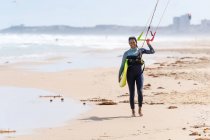 Sportswoman in muta con aquilone gonfiabile passeggiare sulla riva sabbiosa guardando la fotocamera contro l'oceano tempestoso — Foto stock