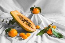 Half of pawpaw with seeds between tangerines and cumquats with leaves on white crumpled cloth — Foto stock