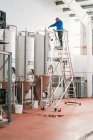 Инженер-мужчина по лестнице заливает жидкость в бак из нержавеющей стали во время работы на пивоваренном заводе — стоковое фото