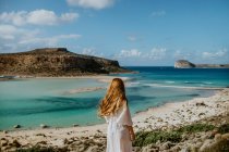 Анонимная женщина в белой макси-пляжной одежде, стоящая на каменистой вершине холма и любующаяся живописным видом на пляж Балос и трогательными длинными волосами в солнечный жаркий день — стоковое фото