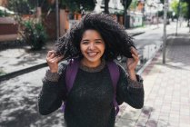 Crop view of delighted ethnic female student with Afro hairstyle and backpack standing on street on sunny day and looking at camera while touching her hair — Photo de stock