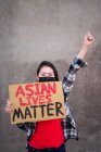 Femme ethnique avec masque et plaque en carton avec inscription Asian Lives Matter protestant avec le bras levé dans la rue de la ville et regardant la caméra — Photo de stock