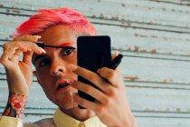 Homem homossexual com piercings e corte de cabelo moderno aplicando rímel em pestanas com aplicador contra celular — Fotografia de Stock