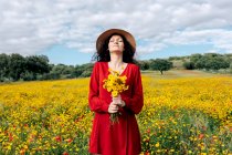 Жінка в капелюсі з закритими очима тримає квітучі жовті квіти в сільській місцевості під хмарним небом — стокове фото