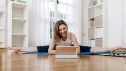 Lächeln flexible Frau praktiziert Yoga in Upavista Konasana, während sie auf Matte sitzt und Online-Video-Tutorial auf dem Tablet zu Hause anschaut — Stockfoto