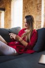 Weibliche Remote-Mitarbeiter SMS auf dem Handy, während sie auf der Couch gegen Tablet in Loft-Stil Haus sitzen — Stockfoto