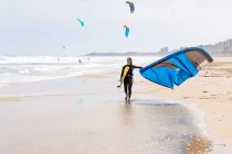 Спортсменка в гідрокостюмі з надувним повітряним змієм, що прогулюється на піщаному березі, дивлячись на штормовий океан — стокове фото