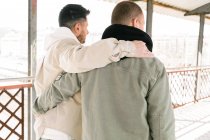 Вид ззаду на невпізнавану пару молодих геїв у стильному одязі, що обіймає один одного, стоячи на мосту в сонячний день — стокове фото