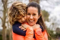 Charming boy embracing smiling mother while sitting in a wooden bench looking away in daylight - foto de stock