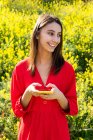 Young content female in red apparel text messaging on cellphone against blossoming plants in sunlight — Stock Photo