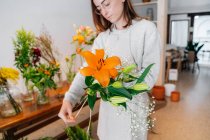 Focada jovem florista étnica feminina em roupas casuais e avental amarrando delicado buquê de lírio laranja e gesso branco na loja floral — Fotografia de Stock