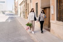 Stylish young female friends in casual clothes and protective masks walking on city street after shopping in grocery during COVID pandemic — Foto stock
