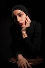 Atractiva joven mujer islámica vestida con traje negro y hijab tocando la cara apoyándose suavemente en la silla en el estudio negro mirando a la cámara - foto de stock