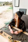 Side view of content female traveler creating handmade accessories while sitting at wooden table in parked truck at seaside — Stock Photo