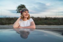 Young female in casual wear and headband with American flag print leaning out of car window while enjoying road trip at sunset — Stock Photo
