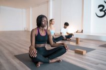 Mujer afroamericana con grupo de diversas personas sentadas en Lotus posan con los ojos cerrados y mediando mientras practican yoga juntos durante la clase en estudio - foto de stock