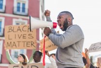 Вид сбоку на африканский американец, кричащий в мегафоне во время протестов черной жизни, имеет значение в городе, стоя в толпе многонациональных демонстрантов — стоковое фото