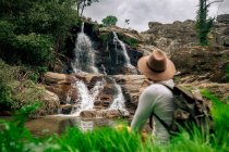 Back view anonymous male backpacker in hat enjoying view of cascade streaming from rough rock in verdant nature — Stock Photo