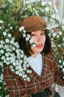 Young gentle ethnic female in checkered wear with beret touching blossoming plant while looking at camera on street - foto de stock
