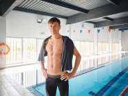 Atleta masculino em roupa de banho com toalha olhando para a câmera contra a piscina após o treinamento — Fotografia de Stock