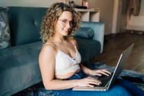 Side view of young self employed woman in casual clothes and eyeglasses sitting on the floor looking at camera near comfortable sofa and working remotely on laptop — Photo de stock