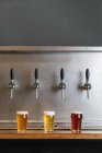 Different types of beer with foam in glass jugs against row of taps in bar on gray background — Stock Photo