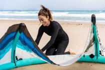 Жінка-кіттер у гідрокостюмі встановлює надувний повітряний змій на піщаному узбережжі океану з рюкзаком та спорядженням на дошці для кіоску — стокове фото