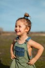 Délicieuse adorable petite fille en salopette debout avec les mains sur la taille dans la prairie et regardant loin — Photo de stock