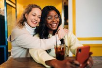 Восхитительные многорасовые женщины лучшие друзья обнимаются в кафе и фотографируются на смартфоне, наслаждаясь выходными вместе — стоковое фото