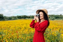 Mulher sorridente de chapéu tirando foto na câmera vintage no prado sob céu nublado — Fotografia de Stock