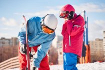 Ajuste o pai e a criança vestindo roupas esportivas quentes e capacetes colocando esquis enquanto estão em pé na colina nevada no subúrbio no inverno ensolarado — Fotografia de Stock