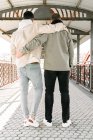 Back view of unrecognizable young gay couple in stylish clothes hugging each other while standing on bridge on sunny day - foto de stock