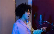 Cantante negra interpretando canción contra micrófono con filtro pop mientras está de pie con la mano en la cadera y los ojos cerrados en el estudio de sonido - foto de stock