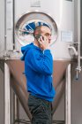 Вид збоку чоловічий підприємець говорить на мобільний телефон проти посудин з нержавіючої сталі на мокрій підлозі на заводі пива — стокове фото