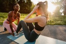 Vista laterale di atleta donna sottile che fa scricchiolii addominali con l'aiuto di sportiva nera durante l'allenamento al tramonto nel parco — Foto stock